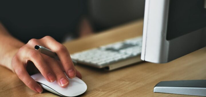 A hand using a wireless mouse at a modern desk setup with a computer and keyboard.