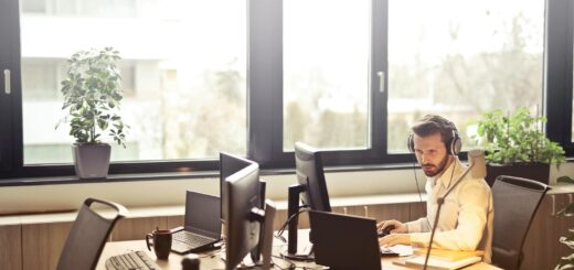 A businessman sits at a desk using multiple computers and a headset in a well-lit modern office.